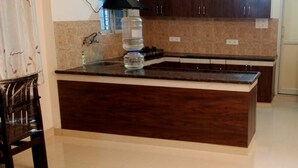 A kitchen with dark wooden cabinets, a granite countertop, a built-in sink, and a refrigerator. - Sri Ramanjaneya Nilayam (Hyderabad)