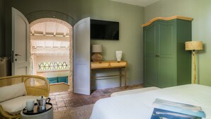 Ein Hotelzimmer mit einem Bett, einem Korbsessel, einem Schreibtisch mit Lampe, einem Kleiderschrank und einem Fernseher.