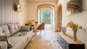 A cozy living room with a sofa, a dining table, and chairs. There is a fireplace, a TV mounted on the wall, and a window with a view of greenery.