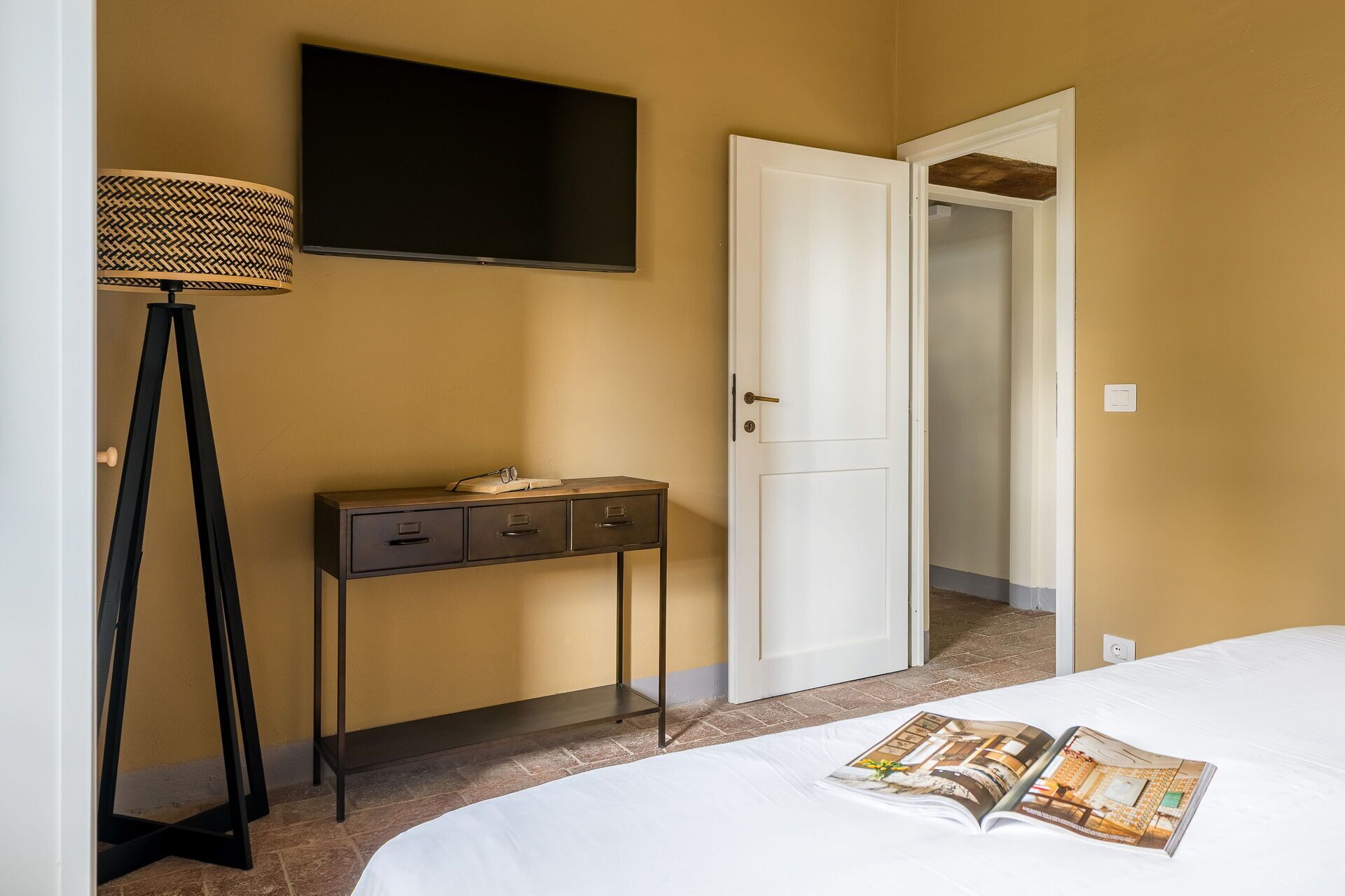 Une chambre d’hôtel avec un lit, une table de chevet avec des tiroirs, une lampe, un téléviseur fixé au mur et une porte donnant sur une autre pièce.
