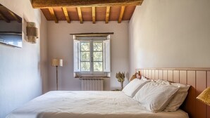 A bedroom with a wooden ceiling, a window with white shutters, a bedside lamp, and a bedside table with a book.