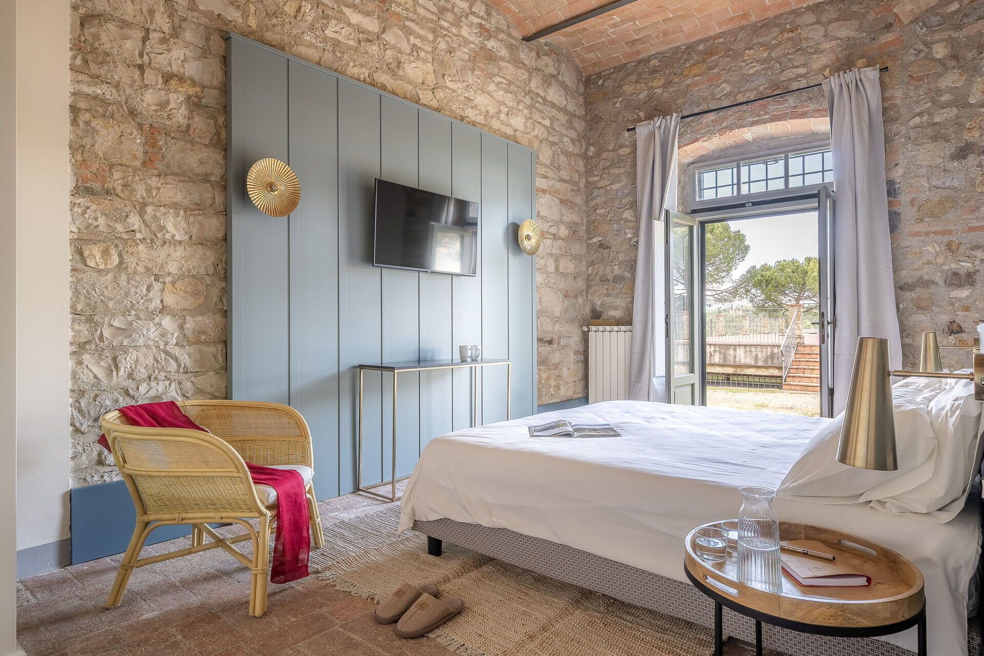 Een slaapkamer met een stenen muur, een bed met een witte spreidek, een rieten stoel, een klein tafeltje met een boek en een glas, en een televisie aan de muur.