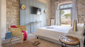 A bedroom with a stone wall, a bed with a white bedspread, a wicker chair, a small table with a book and a glass, and a TV on the wall.
