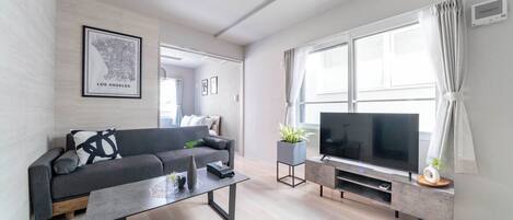 Un salon moderne avec un canapé gris, une table basse, un téléviseur à écran plat et une grande fenêtre avec des rideaux.