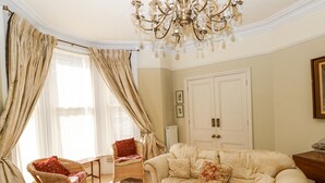 Ett rum med en beige soffa, en flätad fåtölj och en röd kudde. Det finns en kristallkrona, en vit dörr och ett fönster med gardiner.
