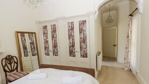 Ett hotellrum med en stor säng, en trästol, en garderob med blommiga gardiner och en välvd dörröppning som leder till ett annat rum.
