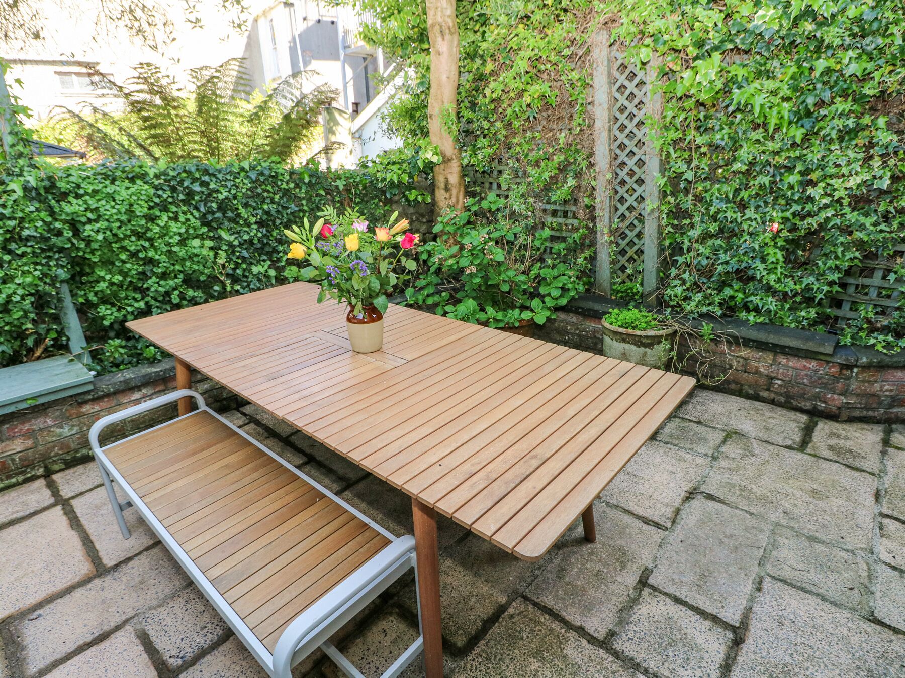 A wooden patio table with a bench, surrounded by lush greenery and a brick wall.