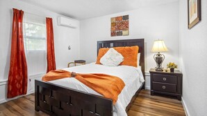 A bedroom with a wooden bed, white bedding, an orange throw, a nightstand with a lamp, and a window with red curtains. - Calm & Spacious Retreat • Family-Ready Escape (Plant City)