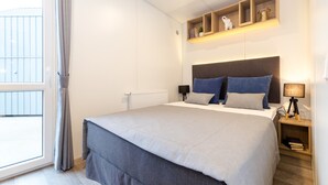 A bedroom with a large bed, a bedside table with a lamp, a shelf with decorative items, and a window with curtains. - Tiny Home 'Ferdl' with Mountain View, Private Garden and Wi-Fi (Untergriesbach)