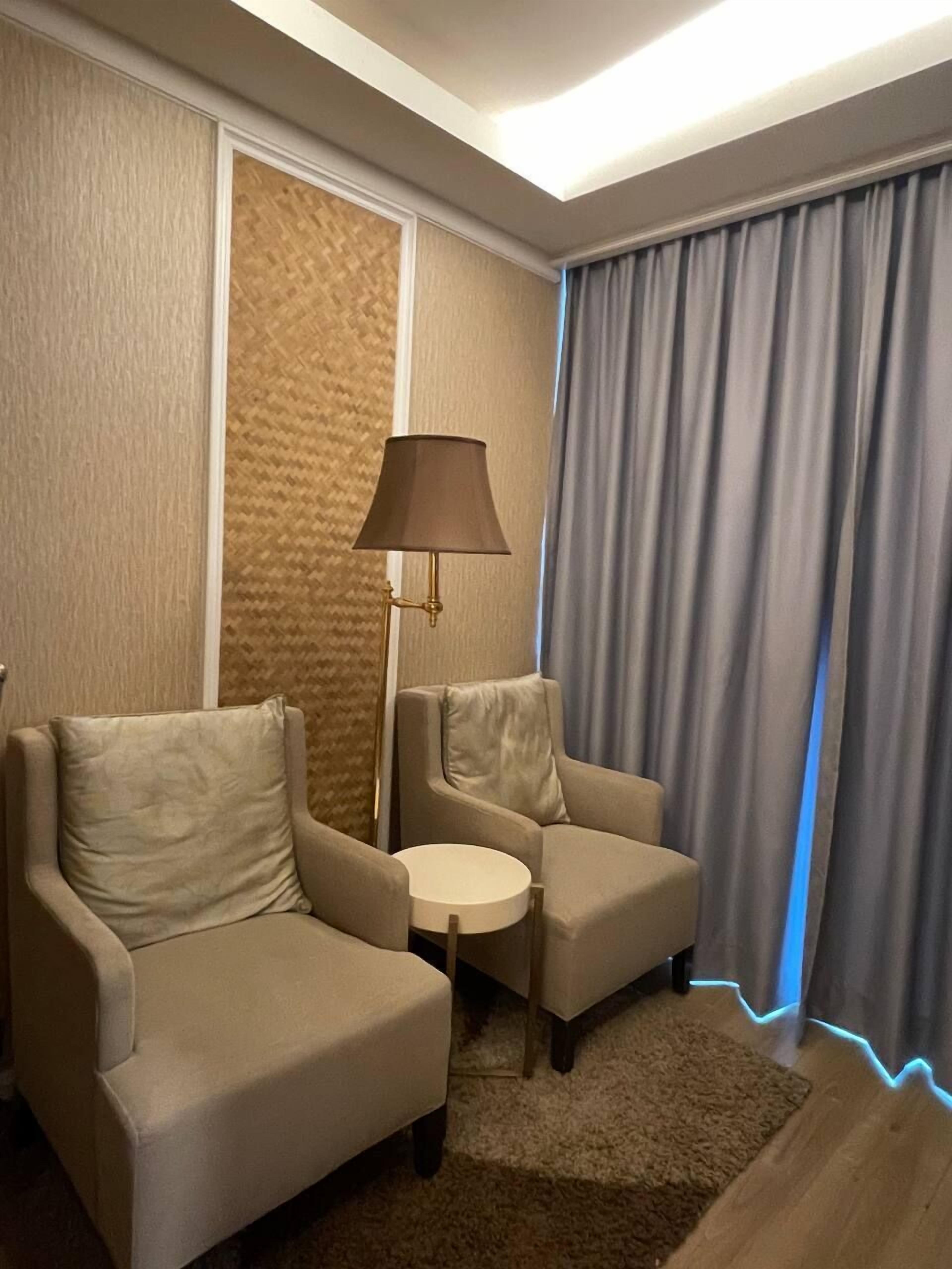 Una habitación con dos sillones, una mesita redonda pequeña, una lámpara de pie y un ventanal con cortinas.