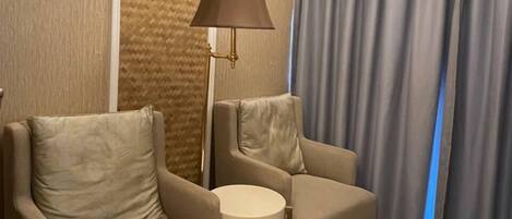 Una habitación con dos sillones, una mesita redonda pequeña, una lámpara de pie y un ventanal con cortinas.