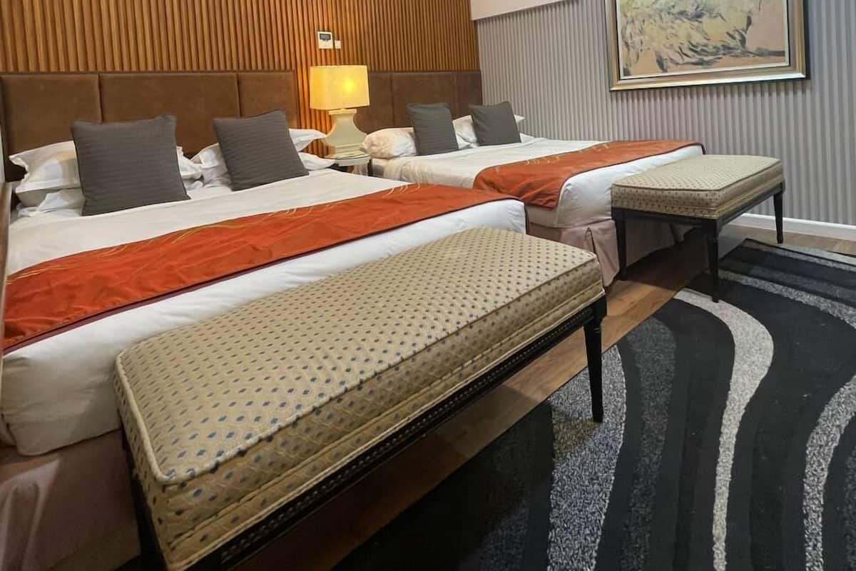 A hotel room with two beds, a wooden sloped ceiling, an air conditioning unit, a painting on the wall, and a black and white patterned rug.