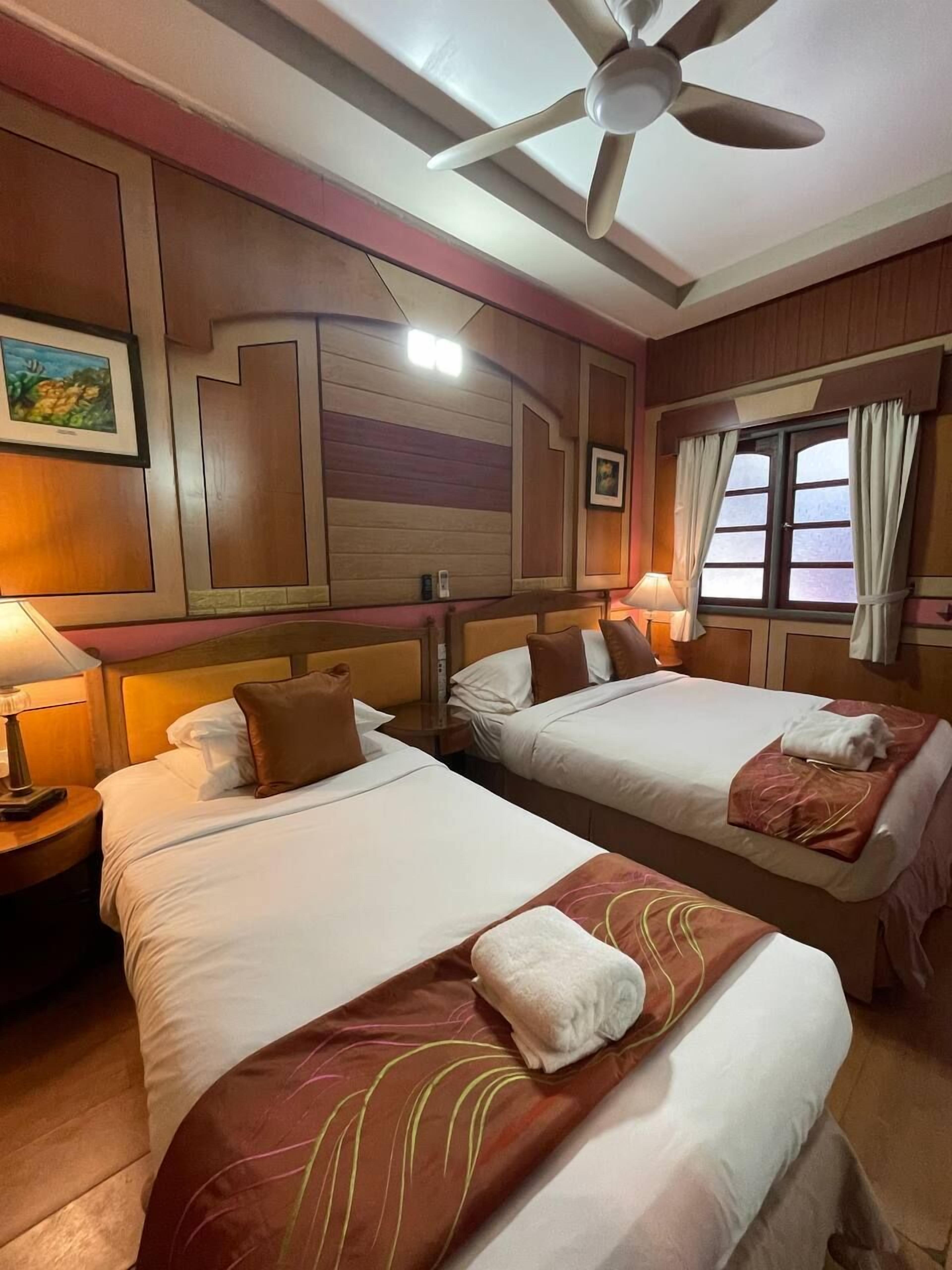A hotel room with two beds, a ceiling fan, a window with curtains, and a painting on the wall.