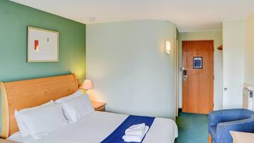 Habitación de hotel con cama, escritorio, silla, puerta y lámpara fijada a la pared.