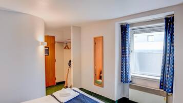 Hotellihuone, jossa on sänky, ikkuna verhoilla, peili ja ovi, joka johtaa toiseen huoneeseen.