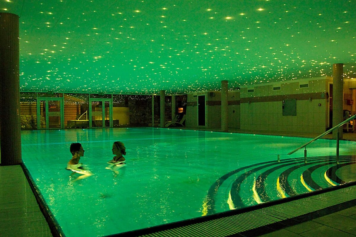 Ein Schwimmbecken mit zwei Personen im Wasser, umgeben von einer mit Fliesen belegten Fläche mit einer Treppe.