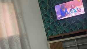 A living room with a TV mounted on a wall with a geometric pattern, a wooden table, and a curtain with a diamond pattern. - Appartement Meublé Moderne - Centre-ville Yaoundé (Air Conditioning, Wi-Fi, Security) (Yaoundé)