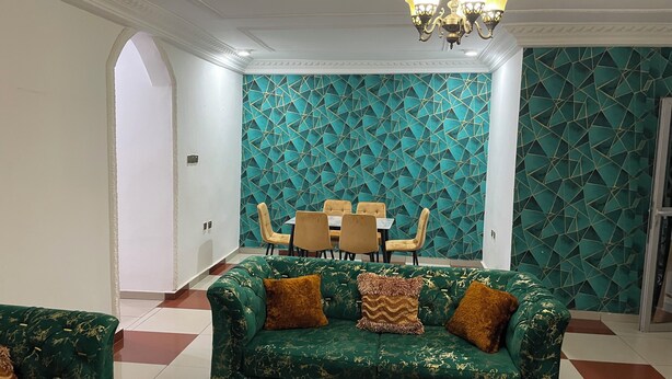 A room with a green sofa, a dining table with chairs, and a patterned wall. - Appartement Meublé Moderne - Centre-ville Yaoundé (Air Conditioning, Wi-Fi, Security) (Yaoundé)