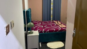A small bedroom with a bed, a bedside table with a lamp, a mirror, and a window with curtains. - Appartement Meublé Moderne - Centre-ville Yaoundé (Air Conditioning, Wi-Fi, Security) (Yaoundé)