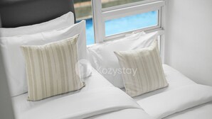 Ein Bett mit weißer Bettwäsche und gestreifter Kissen, ein Fenster mit Blick auf einen Pool und ein grauer Kopfseitenwand.