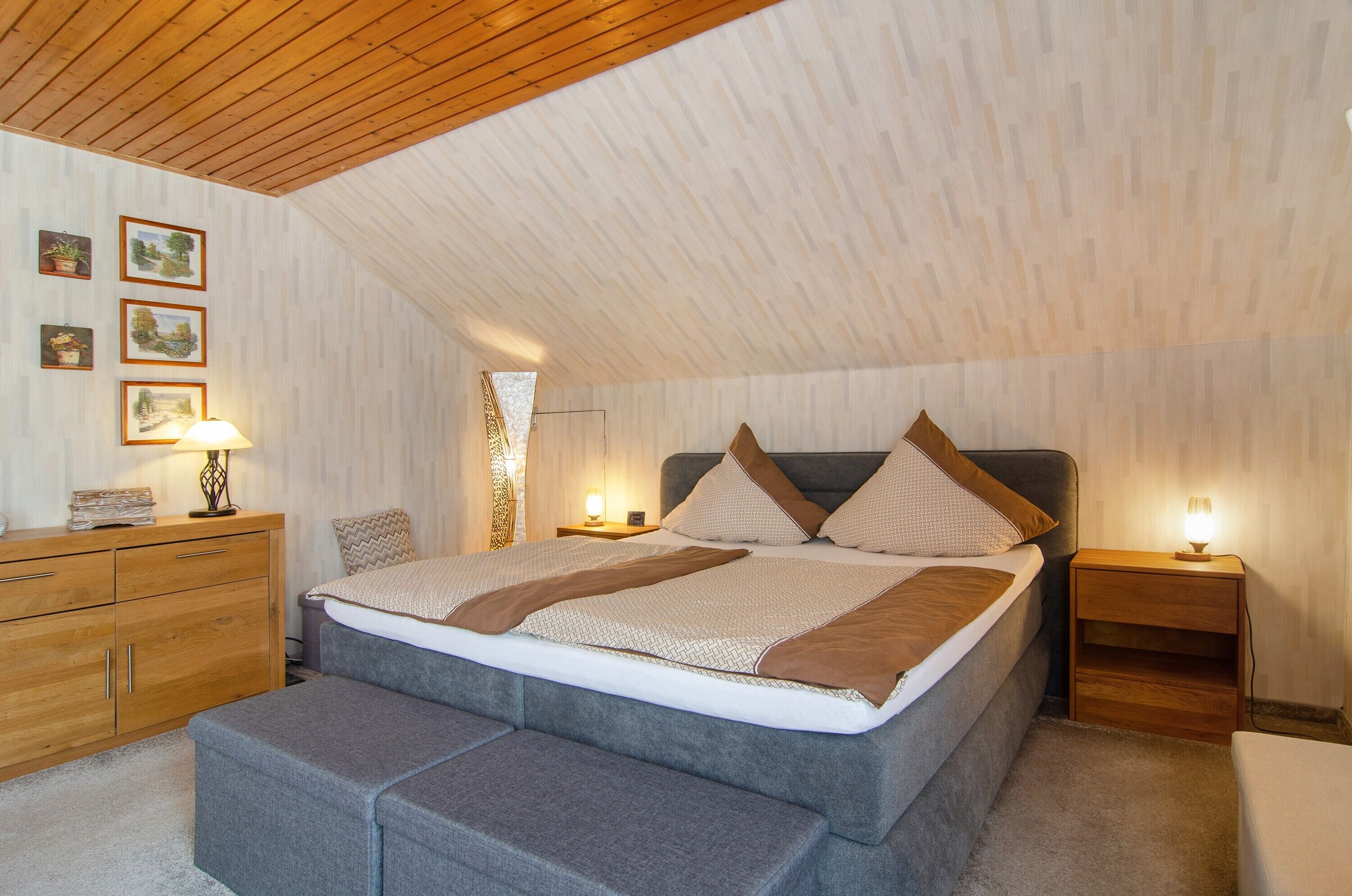 Quarto com teto inclinado, cama com almofadas, mesa de cabeceira com abajur, cómoda de madeira e um banquinho.