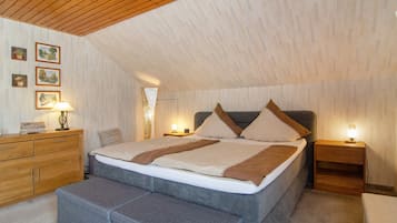 Quarto com teto inclinado, cama com almofadas, mesa de cabeceira com abajur, cómoda de madeira e um banquinho.