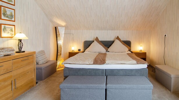 Un dormitorio con techo estilo loft, una cama con muchos almohadones, una mesita de noche con lámpara y un pequeño banco.