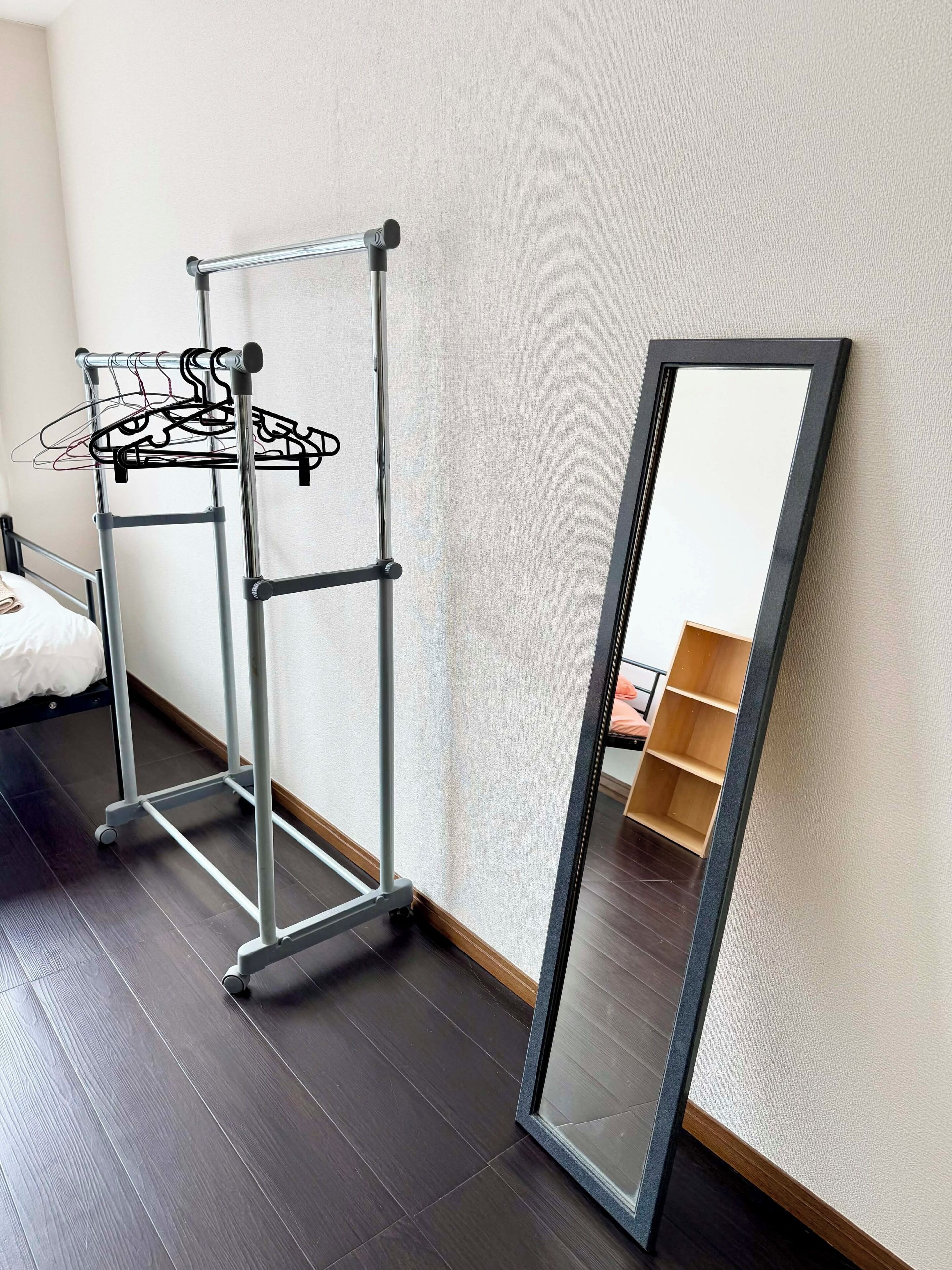 A room with a metal drying rack against the wall, a large mirror leaning against the wall, and a bed with a white duvet.