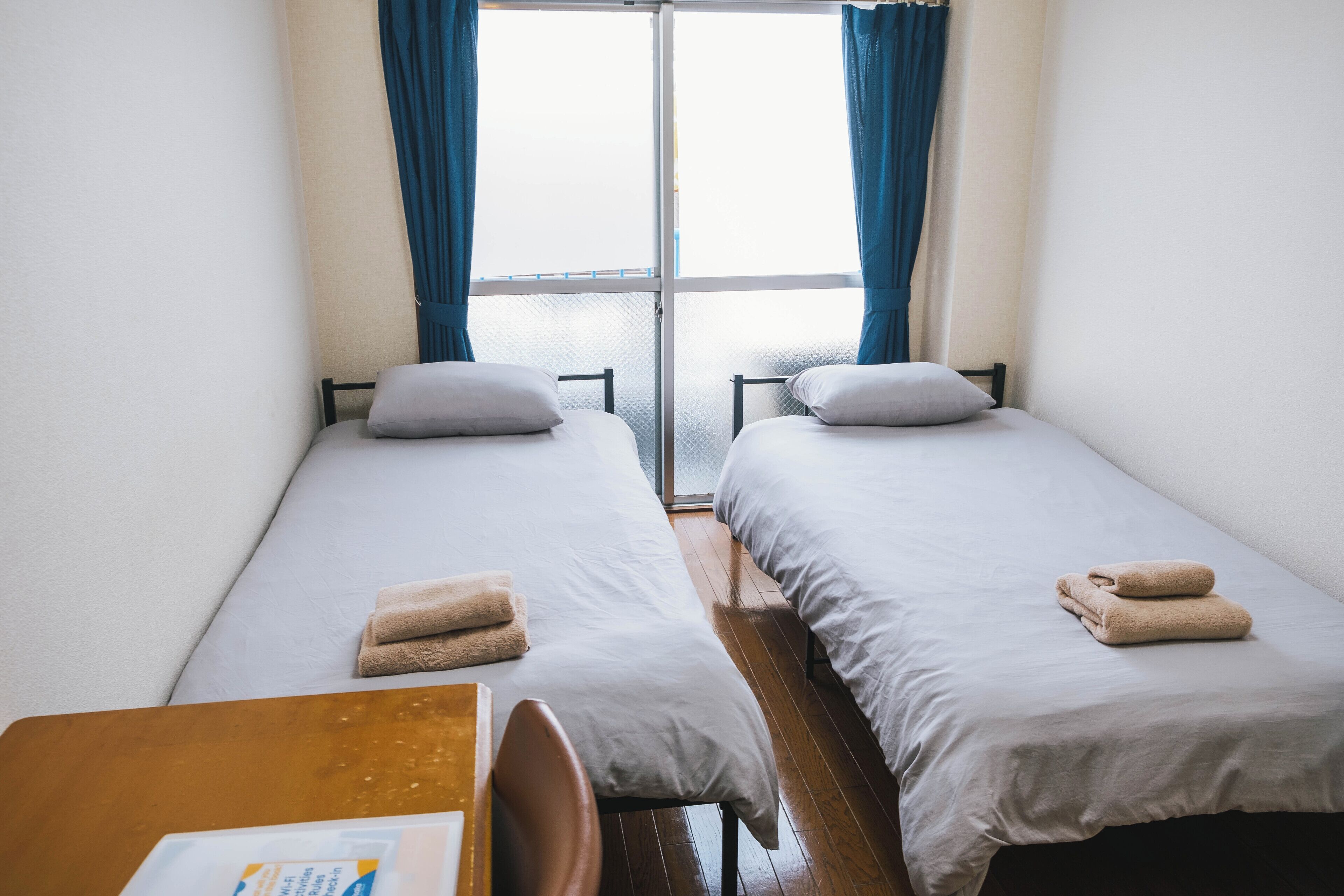Una habitación con dos camas individuales, una mesa de madera y un ventanal con cortinas.