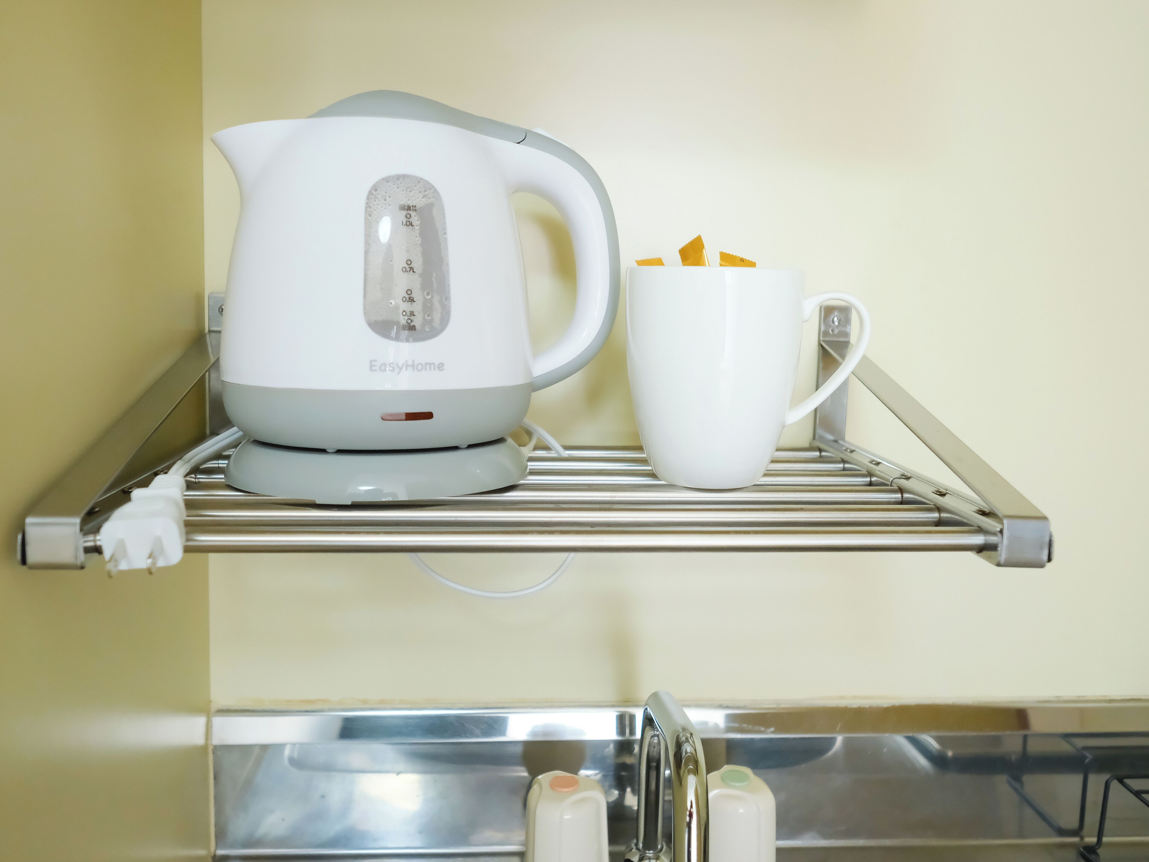 Ein Wasserkocher auf einem Metallregal, neben ihm eine weiße Tasse.