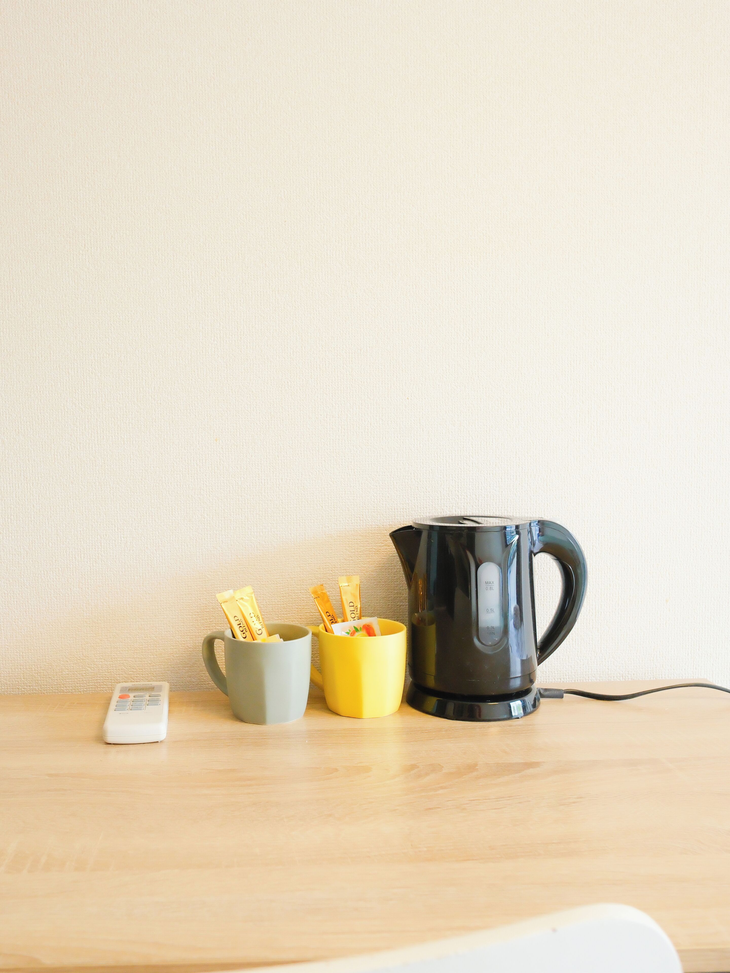 Une table en bois, sur laquelle se trouvent une bouilloire électrique noire, une tasse grise et une tasse jaune contenant des sachets.