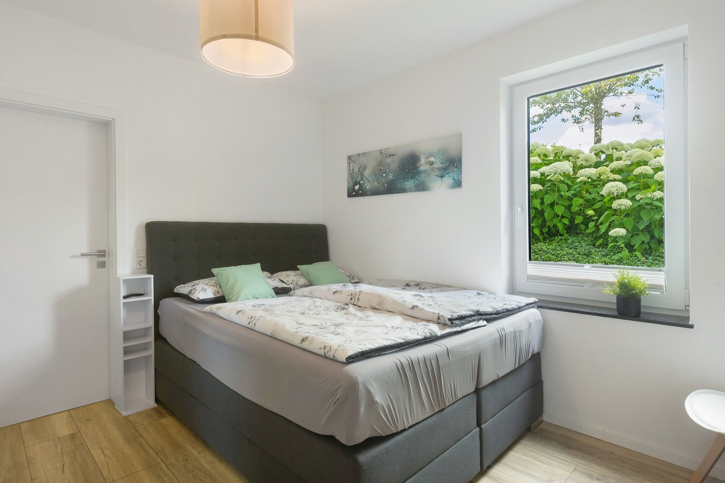 Ein Schlafzimmer mit Bett, einem Fenster mit Blick auf Grünflächen, einem Wandregal und einer Deckenleuchte.