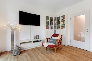 Un salon comprenant un téléviseur à écran plat, une bibliothèque, un fauteuil rouge avec un coussin, une console blanche et un lampadaire.