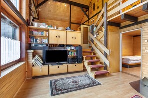 A wooden cabin interior with a TV, bookshelves, a staircase, and a bed.