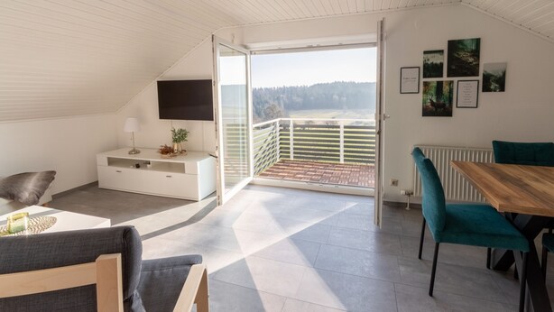 Un dormitorio tipo loft moderno con un amplio ventanal corredera que da a un balcón, una mesa de comedor de madera y un televisor montado en la pared.