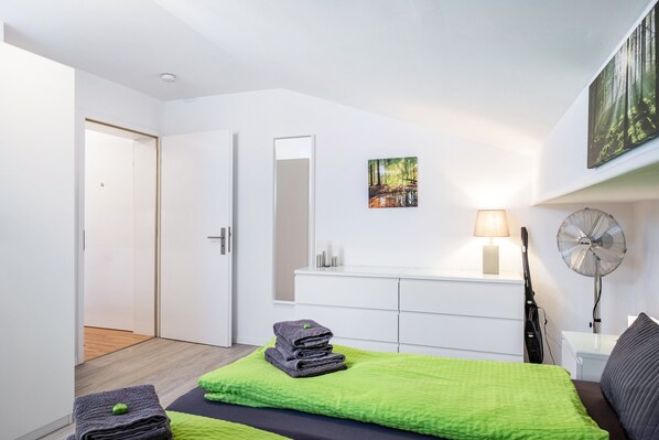 Een moderne slaapkamer met een bed, beddengoed in groen en zwart, een nachtkastje met een lamp, een ventilator en een aan de muur hangend schilderij.
