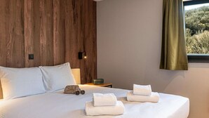A neatly made bed with white linens and towels, a wooden headboard, a bedside table with a lamp, and a window with a view of trees.
