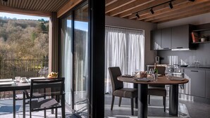 A modern kitchen with a dining area, large windows, and a view of a forested hillside.