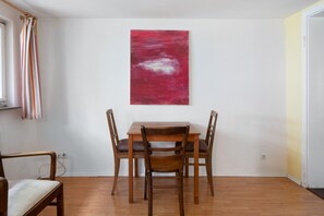 A dining area with a wooden table and chairs, a red abstract painting on the wall, and a window with curtains.