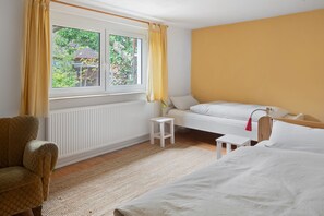 A room with a bed, a small white table, a chair, a window with curtains, and a view of greenery outside.
