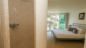 Een slaapkamer met een bed, een douchegedeelte met betegelde wanden en een balkon met uitzicht op bomen.