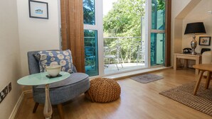 Een moderne woonkamer met een ronde tafel, een blauwe fauteuil, een rieten poef en uitzicht op een balkon met tuinmeubilair.