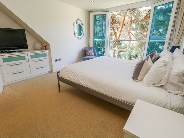 Un dormitorio con cama, televisor sobre un mueble, cómoda y ventana con vista a árboles.