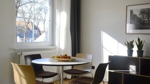 Un comedor con una mesa redonda, sillas, un cuenco de fruta, una ventana con cortinas y un cuadro en la pared.