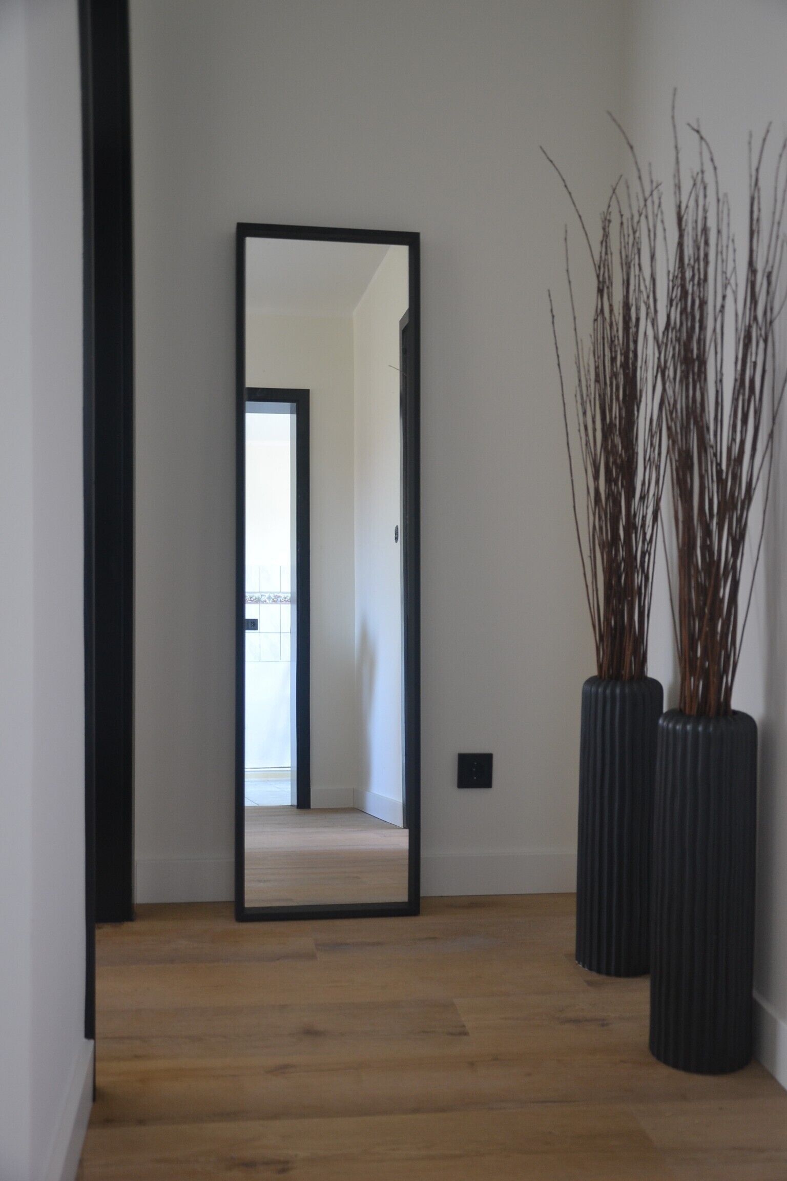Een hal met een spiegel die een kamer weerspiegelt, houten vloeren en twee hoge zwarte vazen met takken.