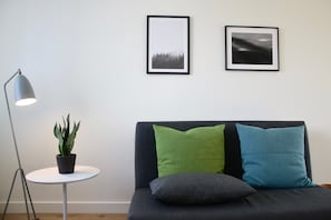 Una habitación con un sofá, dos almohadas, una pequeña mesa con una planta y dos cuadros en la pared.