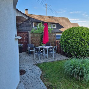 Une terrasse avec une table et des chaises, un parasol rouge et une haie.