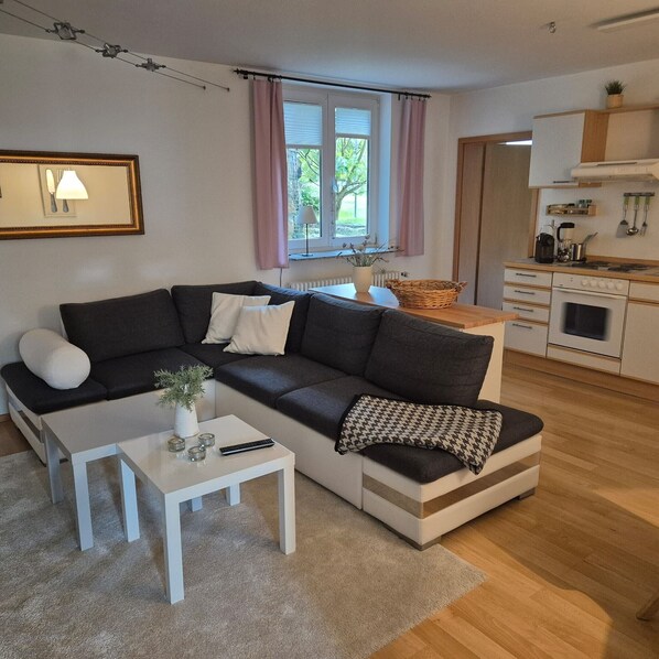 Un salon moderne avec un canapé d’angle, une petite table blanche et un coin cuisine équipé d’une cuisinière et de placards.