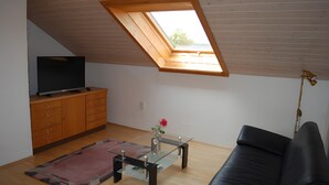 Ein Zimmer mit schrägem Dach, einem Fernseher auf einer Holzkommode, einem Glastisch mit einer Vase, einem schwarzen Ledersofa und einem Holzboden.