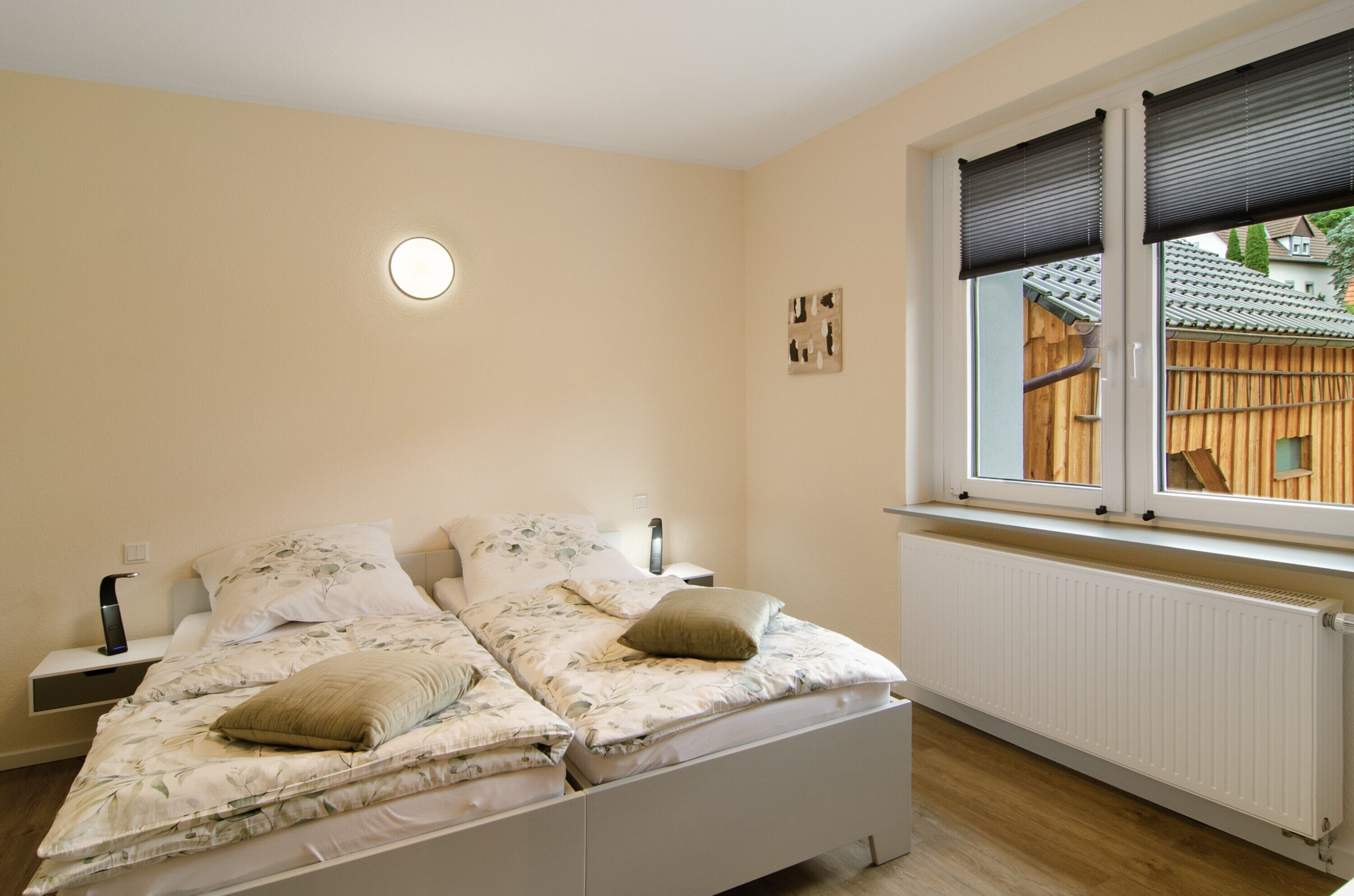 Ein Schlafzimmer mit Bett, Nachttischen, einem Fenster mit Jalousien, einem Heizkörper und einer Wandleuchte.